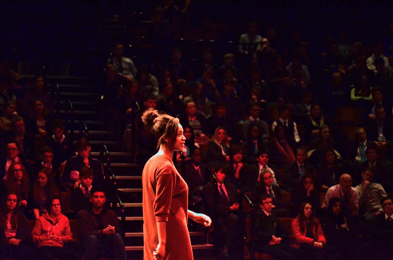 Photo of a presenter, a young black woman, in front of an audience of school students wearing uniform in tiered seating, with red lighting