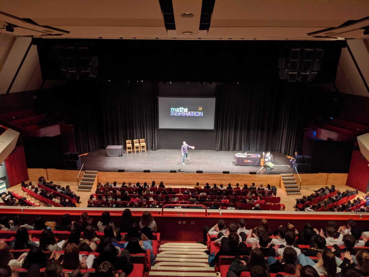 Photo of a theatre stage, with an audience of school students, a slide with the Maths Inspiration logo on screen, and a presenter who is a tall white bald man standing on stage gesturing excitedly