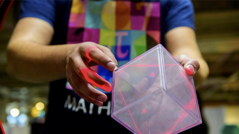 A photograph of a member of the MathsCity explainer team in a branded t-shirt, holding a clear plastic cube in a ring of lasers, showing the cross section of the shape lit up