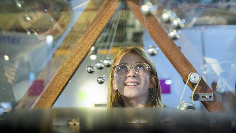 A photograph of a visitor to MathsCity watching a set of pendulums of different lengths swinging through a wooden frame