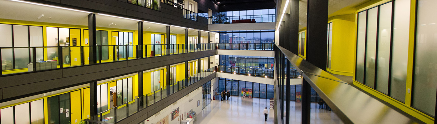 Photograph of the interior of the Alan Turing Building, showing research offices and corridors with yellow walls and a full-height window at the end of the building. A few people are visible standing around and there is a pop-up banner of Alan Turing on the ground floor