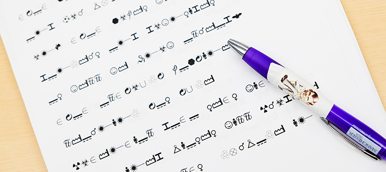 Promotional image for the event, showing a piece of paper covered in Wingdings symbols with an encoded message, and a purple University of Manchester pen with Alan Turing’s face on it resting nearby