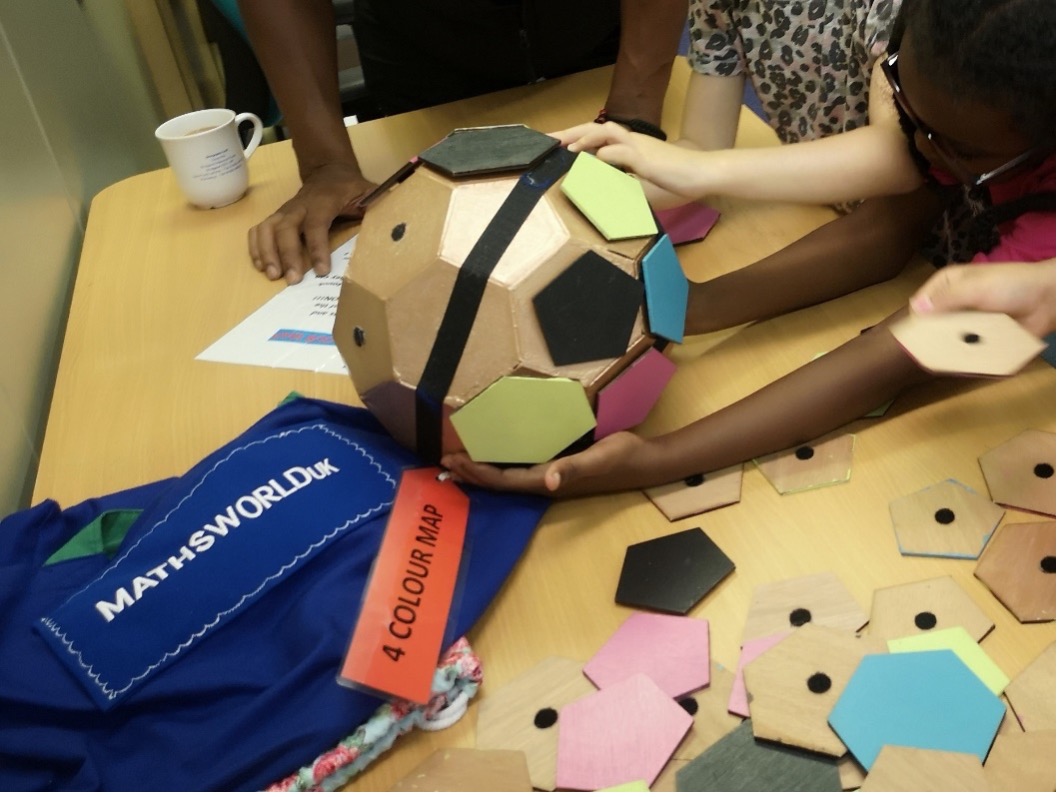 A photograph of children from Gipton Together summer camp standing around a table with their hands on a large wooden truncated icosahedron, which has coloured pieces that can be stuck to each face and is labelled ‘4 COLOUR MAP’