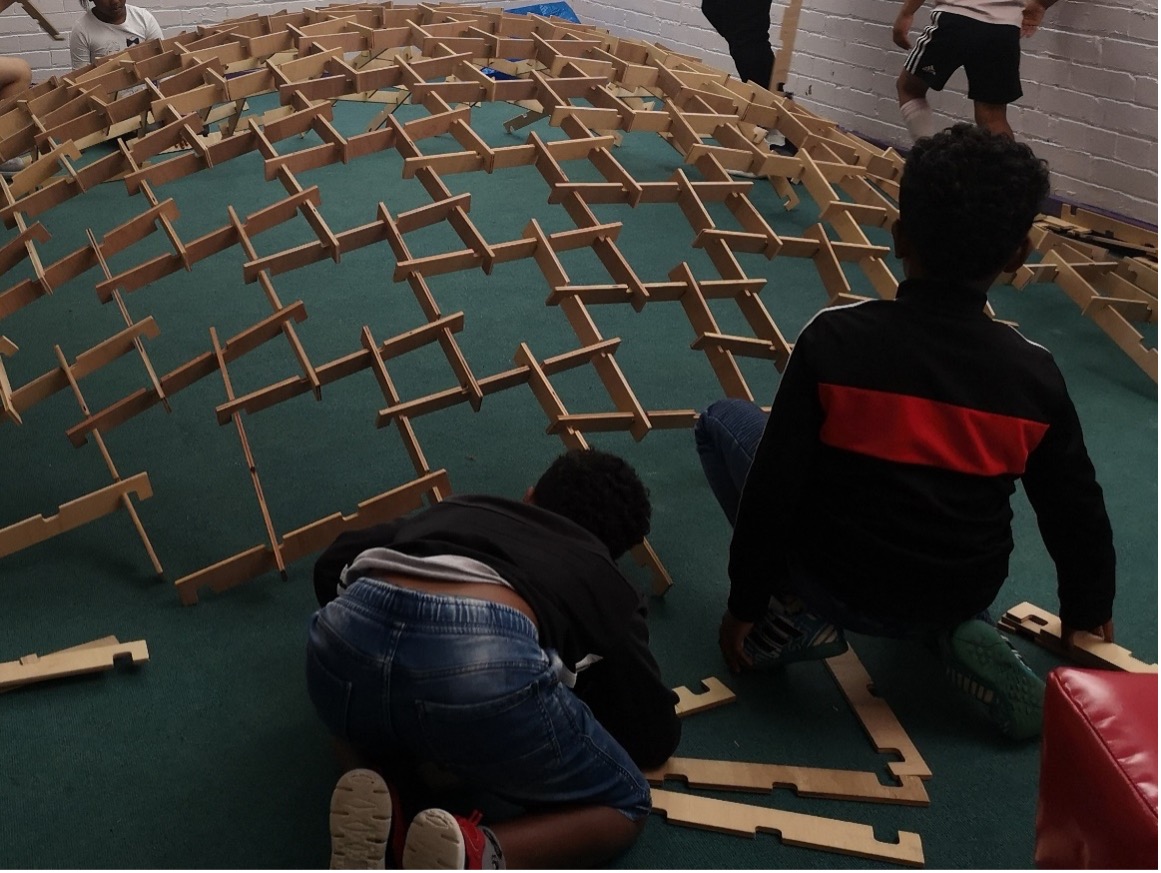 A photograph of two children from Gipton Together summer camp kneeling on the floor, constructing a Leonardo Dome - using strips of wood with notches, hooked together into a shallow dome shape