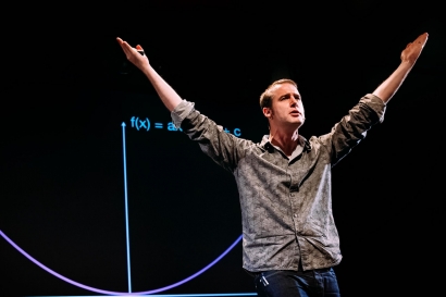 Matt Parker, standing in front of a projector screen with hands in the air