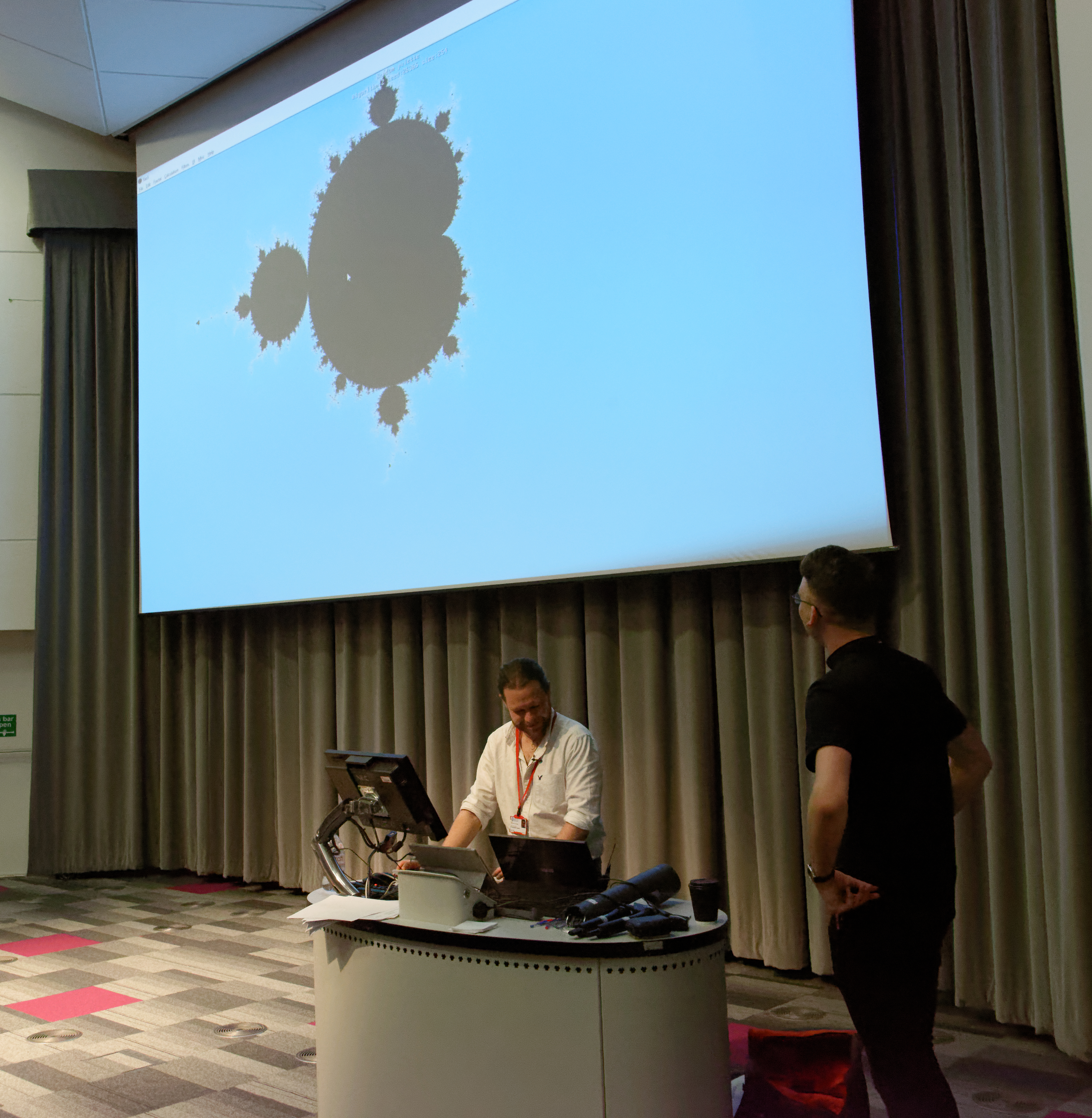 Two men at a lectern in front of a large screen displaying an image of the Mandelbrot set.